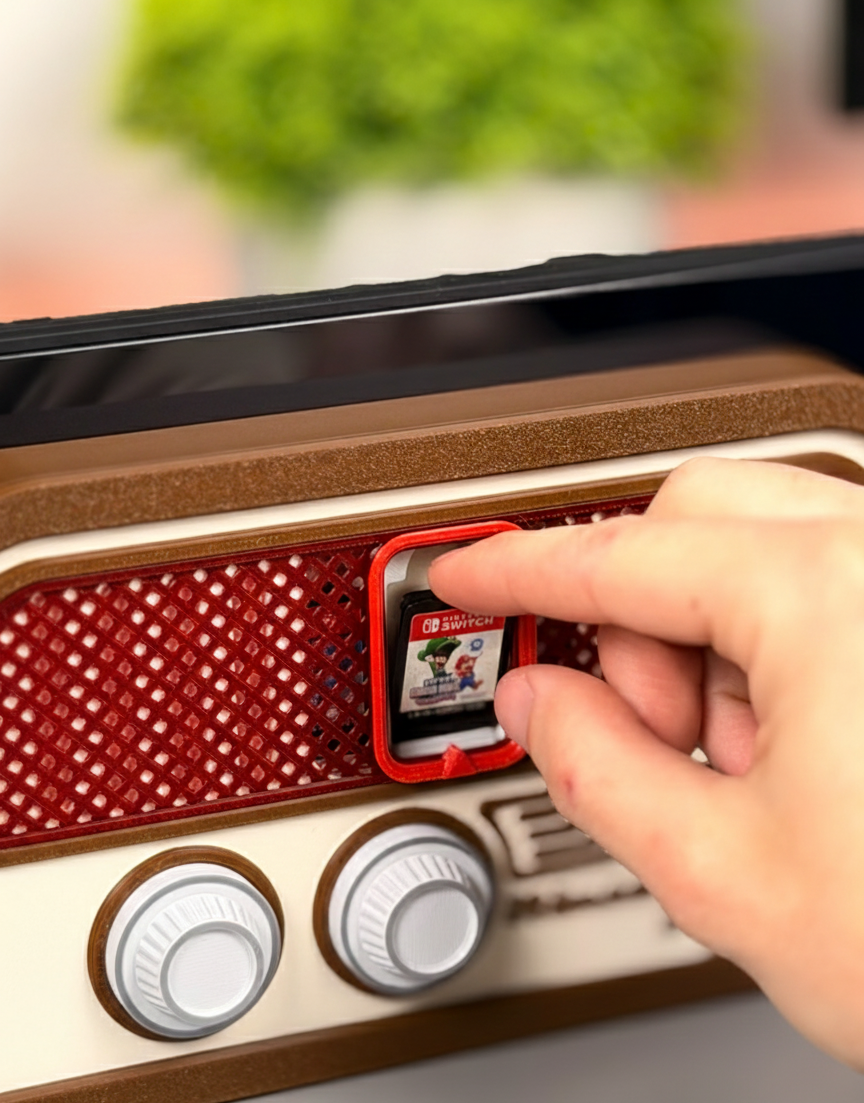 A close-up photograph of a Nintendo Switch console placed in a brown and beige docking stand designed in the style of a vintage radio. A red Nintendo Switch game cartridge (Zelda) is visible in the radio's central screen area, and there are two white control knobs below it. The image is a product shot on a gray desk with a blurred background featuring a green potted plant.