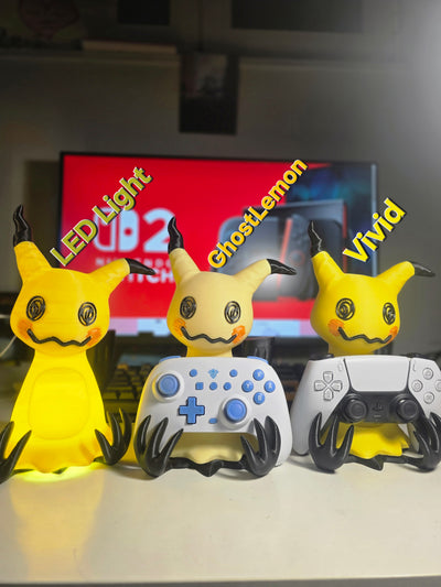 Three Pikachu-themed game controller stands on a white surface in front of a blurred TV. The left one glows with yellow LED light, the middle one is white with blue accents and labeled 'GhostLemon', and the right one is white with black accents and labeled 'Vivid'.