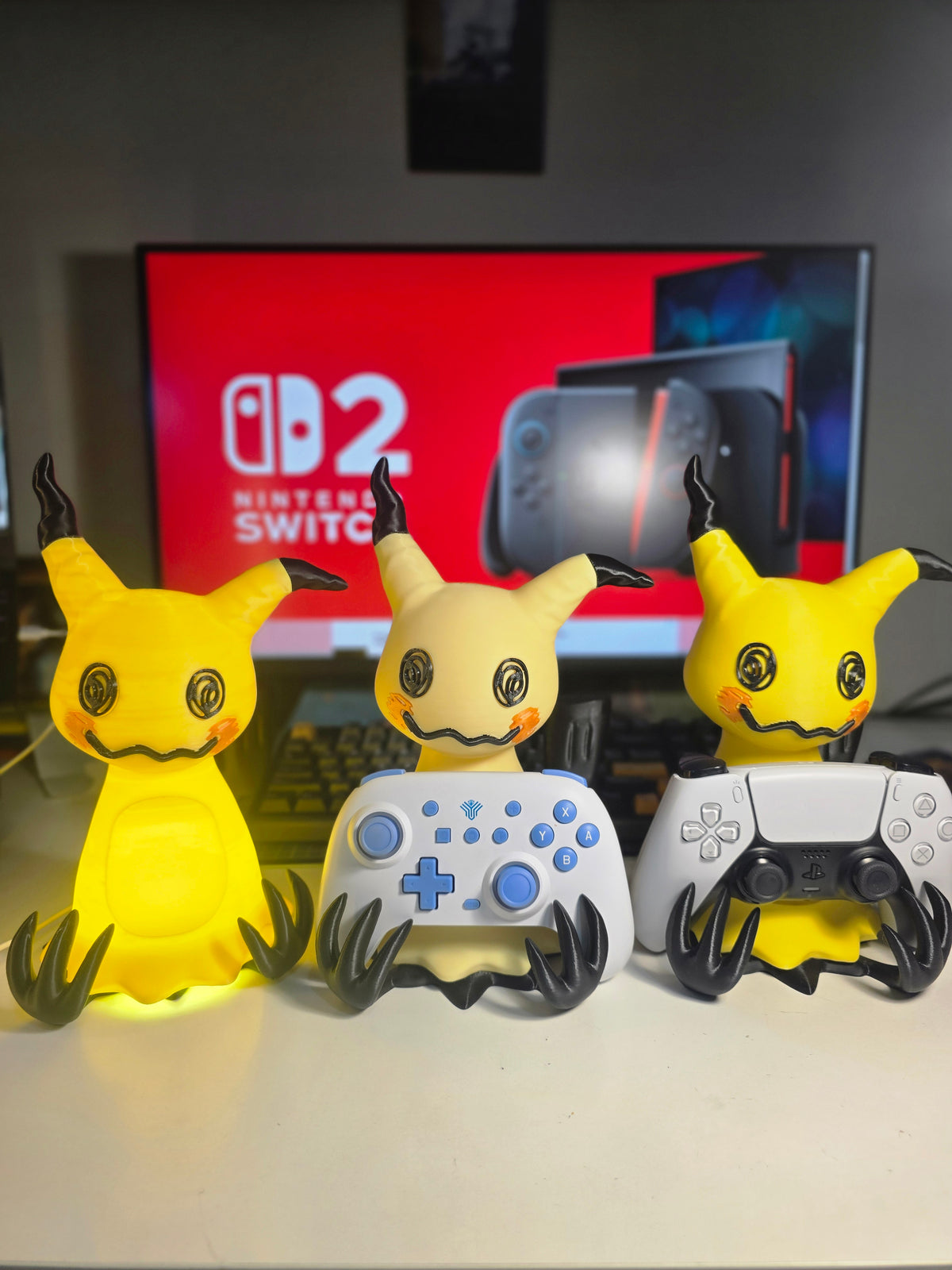 Three Mimikyu figurines in different colors (yellow, cream, and ghost lemon yellow) sitting on a white surface, each holding a game controller (Nintendo Switch, white PS5, and a black PS5). A Nintendo Switch TV screen is visible in the background.